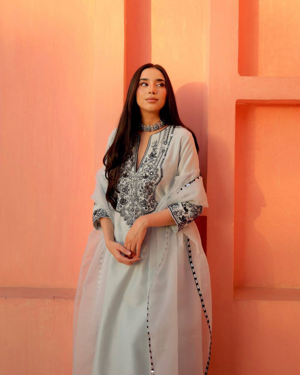 Woman in a light blue traditional outfit with black patterns against an orange wall.